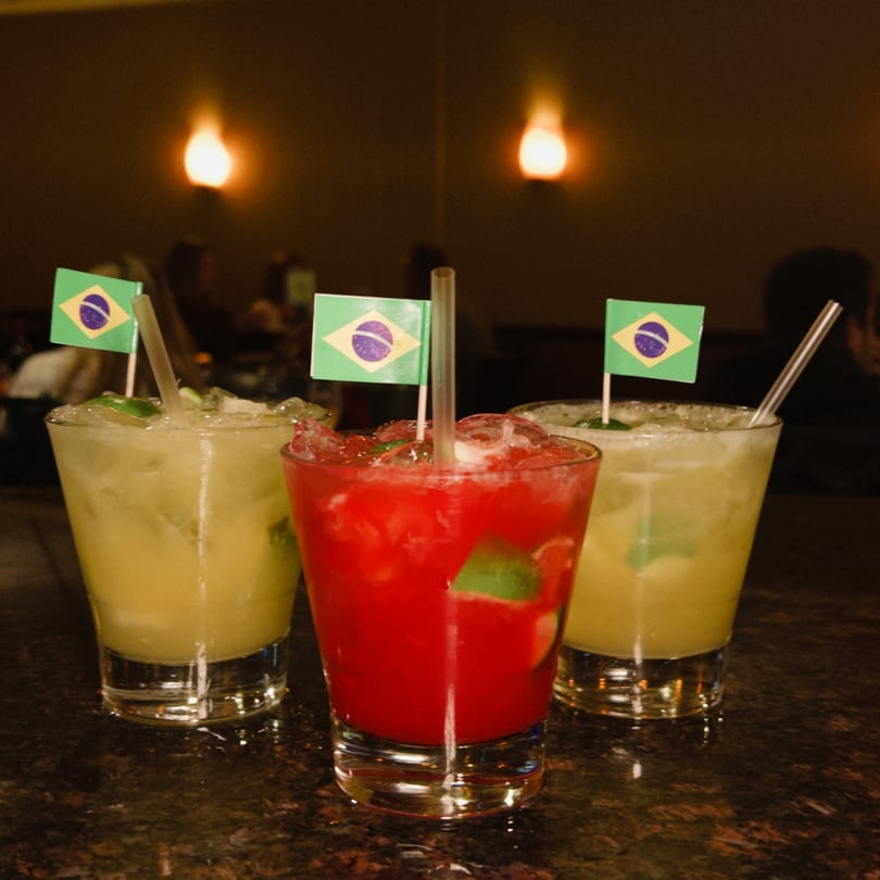 Three Brazilian caipirinha cocktails, two yellow and one red, garnished with lime and Brazilian flags.